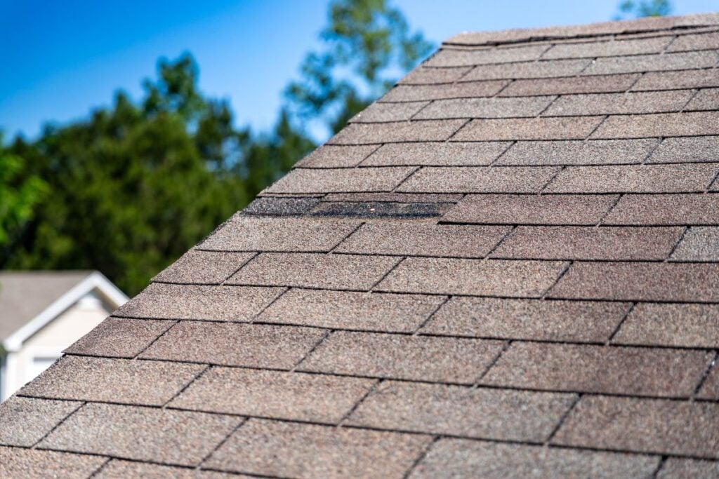 A house roof with asphalt shingles, showing one missing shingle near the center. Trees and a blurred house are visible in the background on a clear, sunny day.