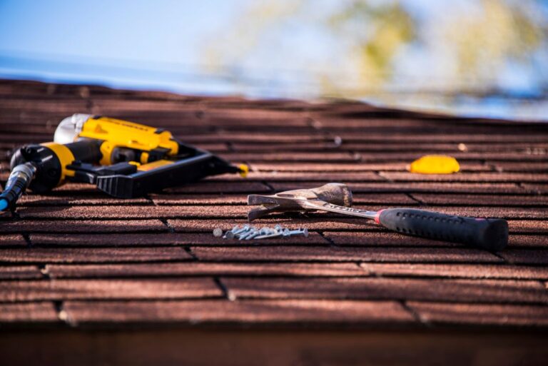 A hammer, nails, and a nail gun lie on a brown shingle roof under a clear sky, with autumn leaves scattered nearby.