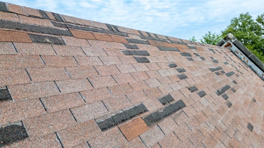 Close-up view of an asphalt shingle roof with a mix of reddish-brown and dark-colored shingles under a partly cloudy sky. Some trees are visible in the background.
