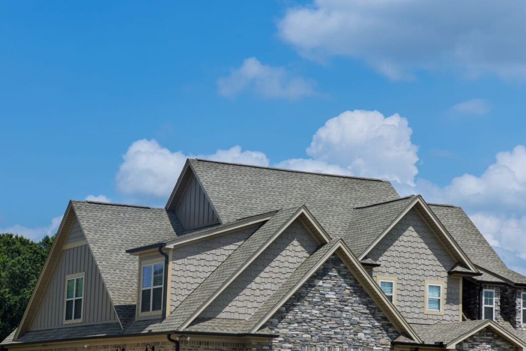 Several peaked rooftops of a modern house with light-colored shingles, stone and siding details, set against a bright blue sky with scattered white clouds.