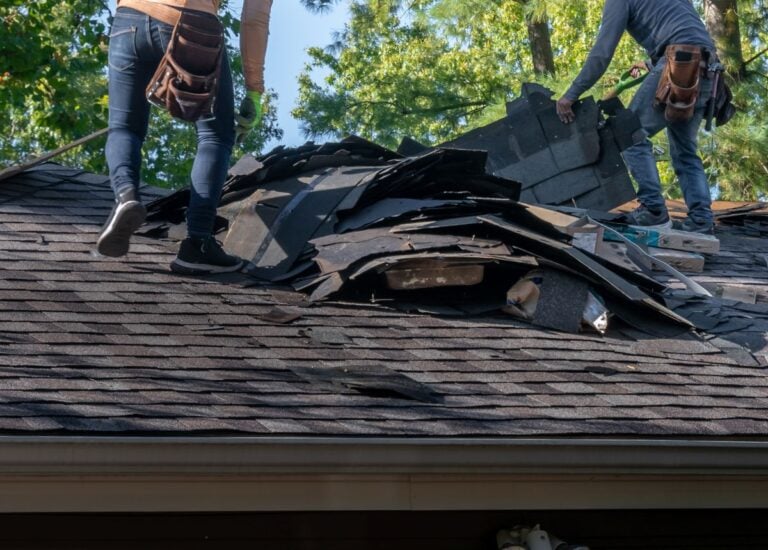 Two workers removing old shingles from a residential roof, piling debris as they prepare for new roofing installation. Trees and a blue sky are visible in the background.
