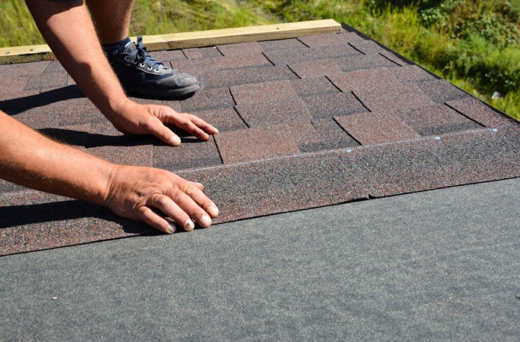 A person in sneakers installs brown asphalt shingles on a roof, pressing down on the edges with both hands. Green grass is visible in the background.