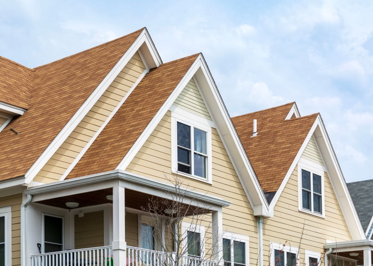 A close-up of a house with beige siding, white trim, multiple gabled roofs with brown shingles, and several windows. A small covered porch and some tree branches are visible in the foreground.