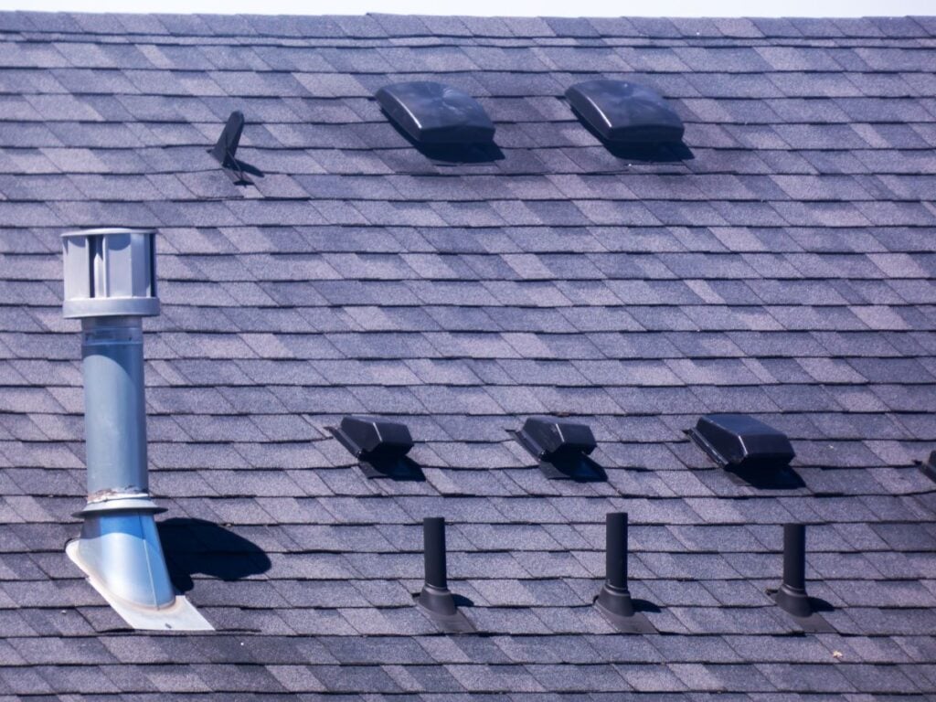 A shingled roof with three metal pipes, several black roof vents, and a large metal exhaust pipe. The sun is shining, casting shadows on the gray shingles.