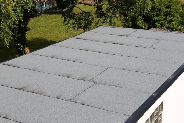 A flat roof covered with light gray roofing material, showing some dark streaks and seams, with green grass and trees visible in the background.