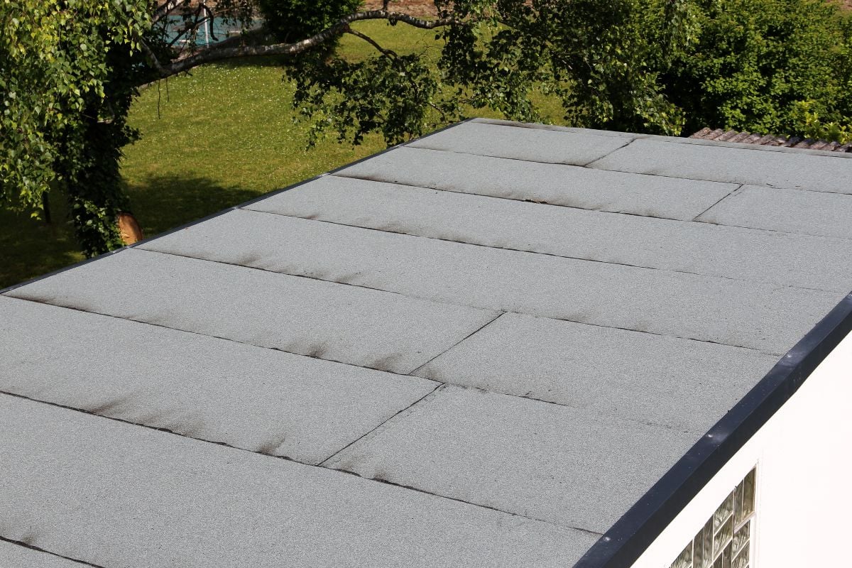 A flat roof covered with light gray roofing material, showing some dark streaks and seams, with green grass and trees visible in the background.