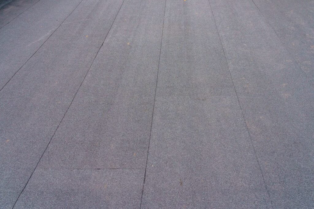 Close-up view of a flat, gray, asphalt or tar paper roof with visible seams and slight texture. The surface appears uniform and is possibly part of a commercial or residential building.