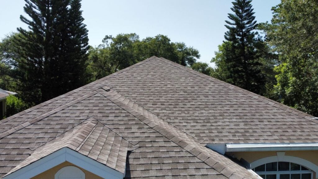 A close-up view of a house rooftop with brown asphalt shingles, surrounded by green trees and a clear blue sky in the background.