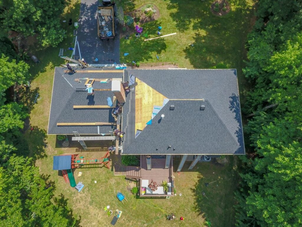 Aerial view of a house with workers repairing the roof. Part of the roof is stripped to the wood. Construction materials and tools are scattered around, and trees and grass surround the house.