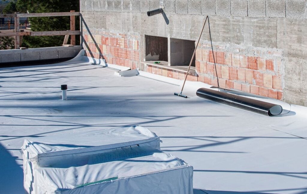 A flat rooftop covered with white material, construction tools, a rolled membrane, a railing, and a partially finished brick and concrete wall in the background.