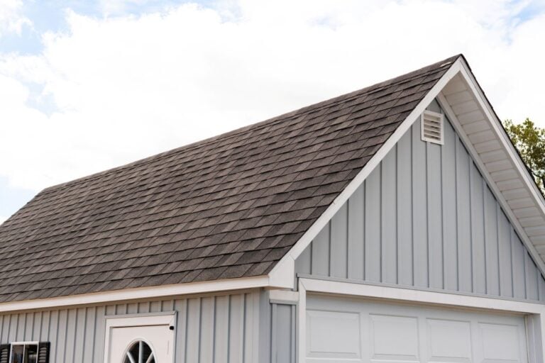 A light blue house with vertical siding and a dark gray shingled gable roof. The white garage door and part of a round window above a door are visible under a partly cloudy sky.