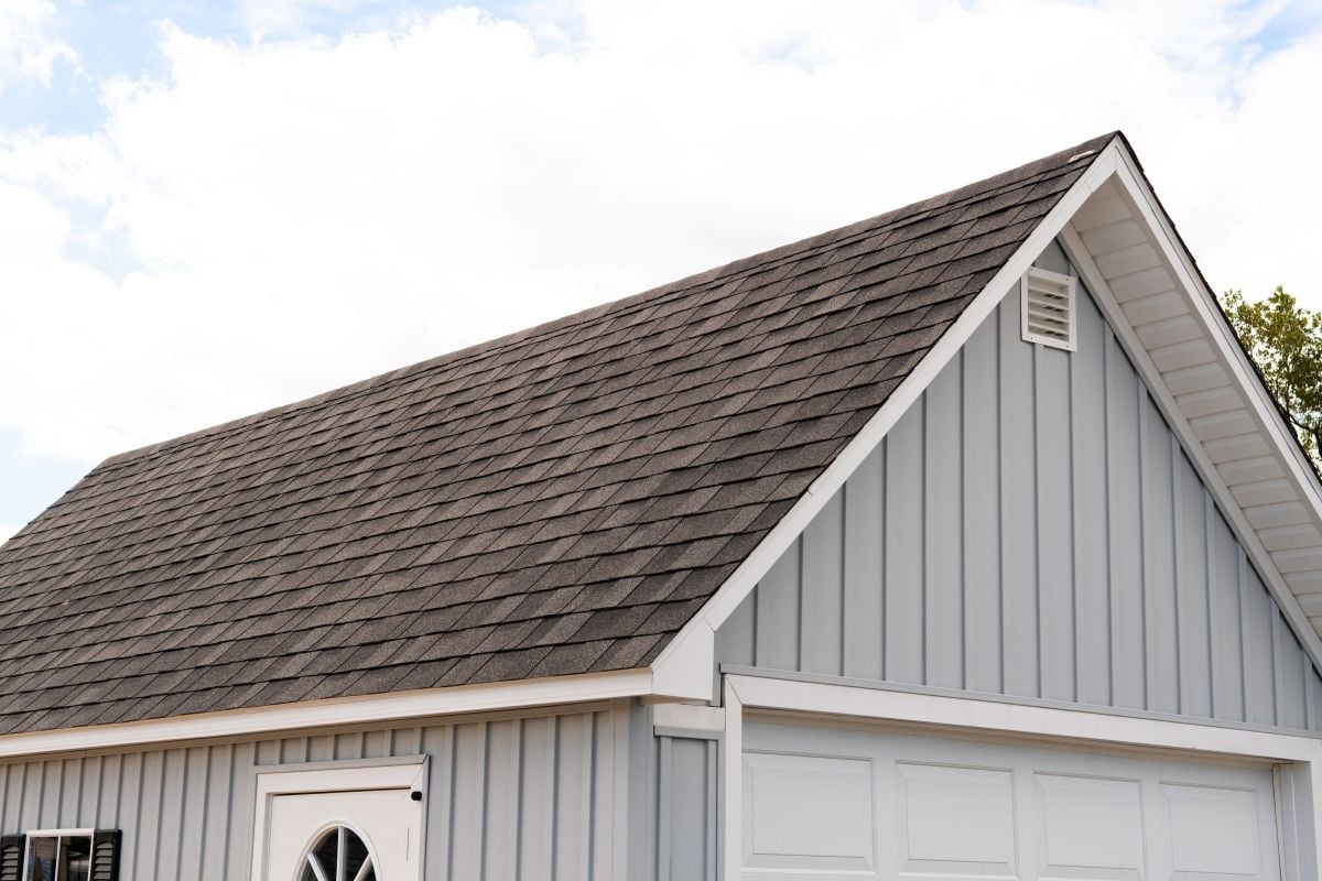 A light blue house with vertical siding and a dark gray shingled gable roof. The white garage door and part of a round window above a door are visible under a partly cloudy sky.