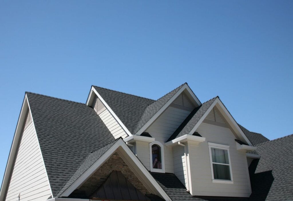 A close-up view of a house with multiple dark gray shingled roof peaks and light-colored siding, set against a clear blue sky.
