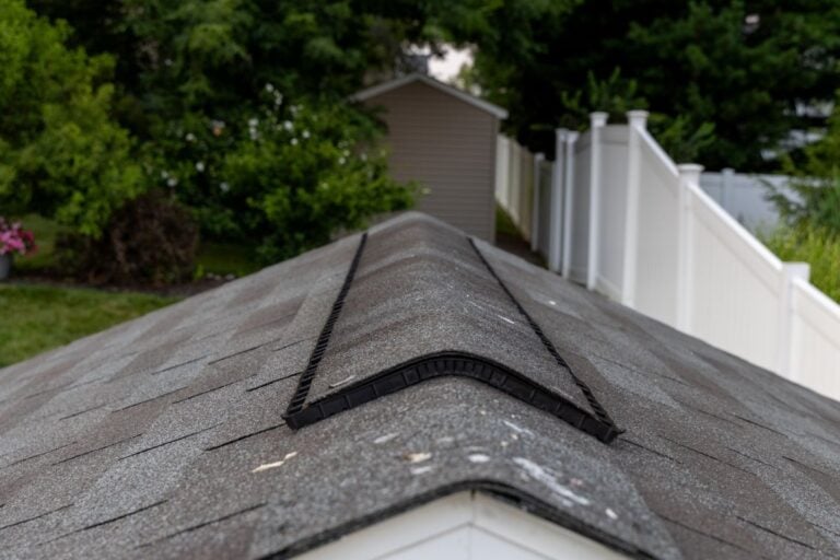 Close-up view of a shingled roof with a ridge vent, set in a green backyard. A white fence runs along the side, leading to a small shed or outbuilding in the background.