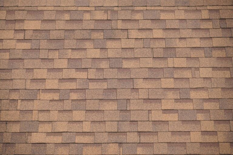 A close-up view of a brown asphalt shingle roof, showing the rectangular pattern and textured surface of the overlapping shingles.