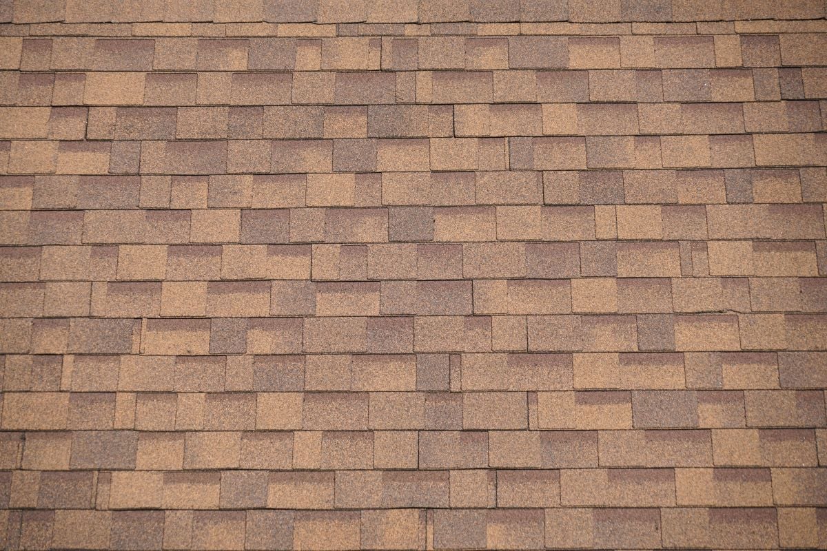 A close-up view of a brown asphalt shingle roof, showing the rectangular pattern and textured surface of the overlapping shingles.