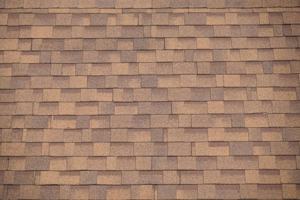 Close-up of an asphalt shingle roof with overlapping brown and tan rectangular shingles in a repeating pattern, showing texture and uniformity.