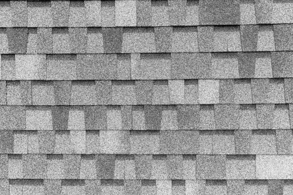 Close-up view of gray asphalt roof shingles arranged in a staggered pattern, showing texture and repeating rectangular shapes with varying shades of gray.