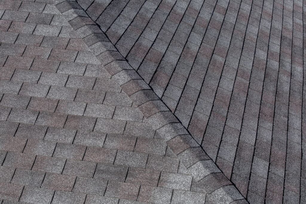 Close-up view of an asphalt shingle roof showing a ridge where two roof sections meet. The shingles are gray and brown, arranged in neat, overlapping rows for weather protection.
