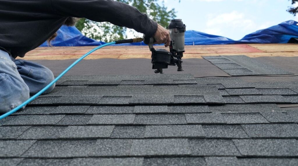 Person using a nail gun to secure asphalt shingles onto a roof, with blue sky and trees in the background.