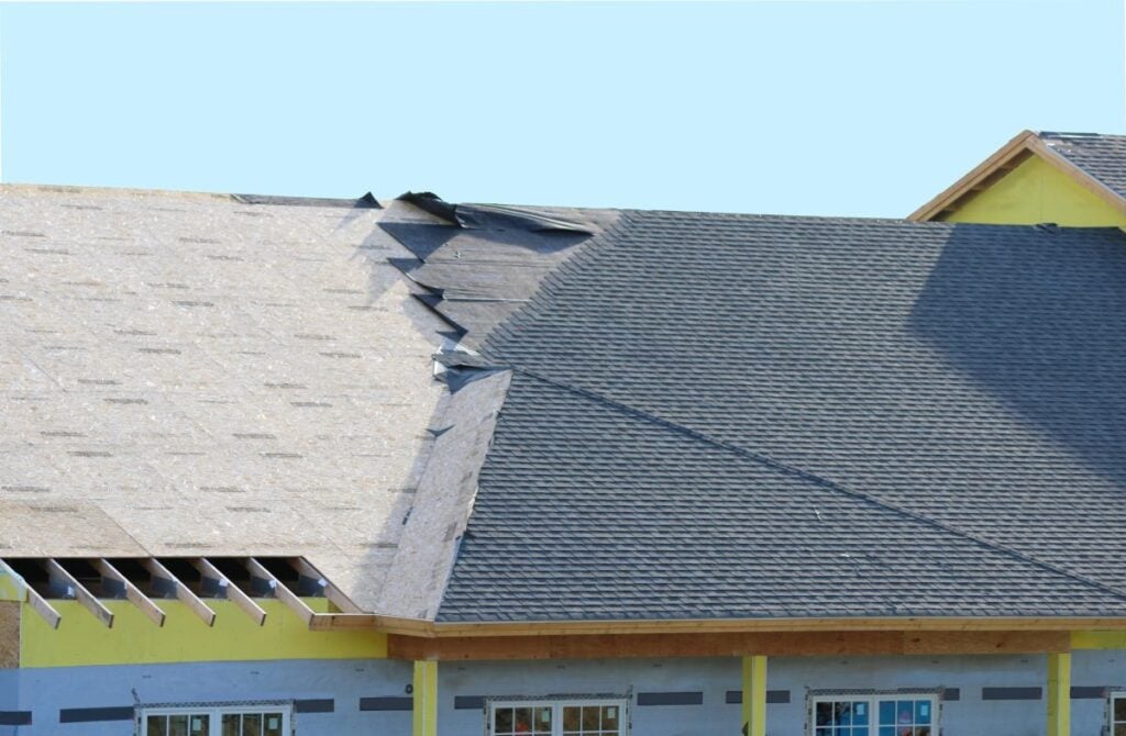 Partially completed house roof showing new shingles on one side and exposed underlayment and wooden structure on the other side, under a clear blue sky.
