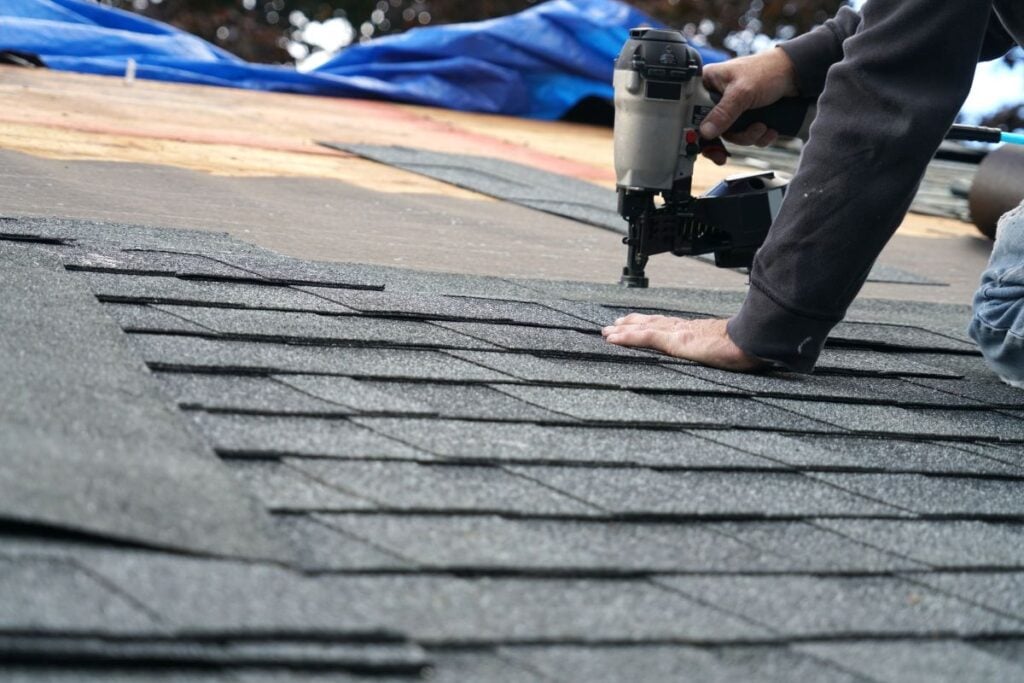 A person installs asphalt shingles on a roof using a nail gun. The person is kneeling, wearing a dark sleeve, and securing the shingles in neat rows.