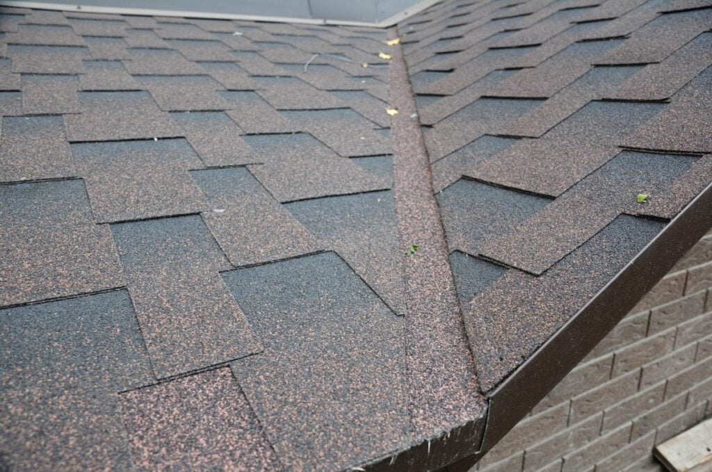 Close-up of a house roof with brown asphalt shingles, showing a clean V-shaped valley where two roof sections meet. Some small debris and leaves are scattered on the shingles.