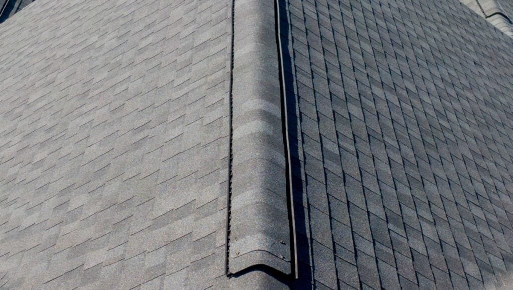 Close-up view of a rooftop with asphalt shingles arranged in a neat, overlapping pattern. A ridge cap runs along the peak of the roof, covering the seam where the two sides meet.