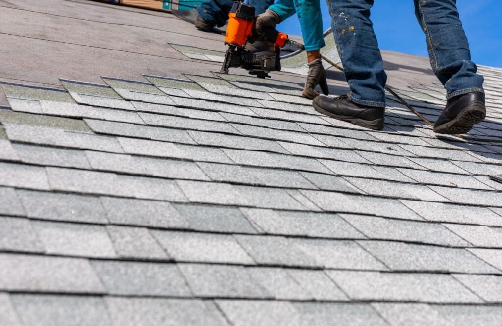 Two workers in jeans and gloves install asphalt shingles on a sloped roof using a nail gun. The roof is partially completed, and the sky is clear and blue.