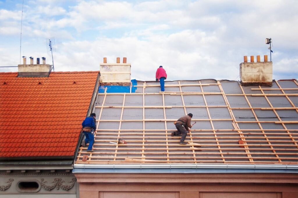 Three people work on a sloped roof installing wooden battens. The roof on the left is finished with red tiles, while the right side is under construction. The sky is partly cloudy.
