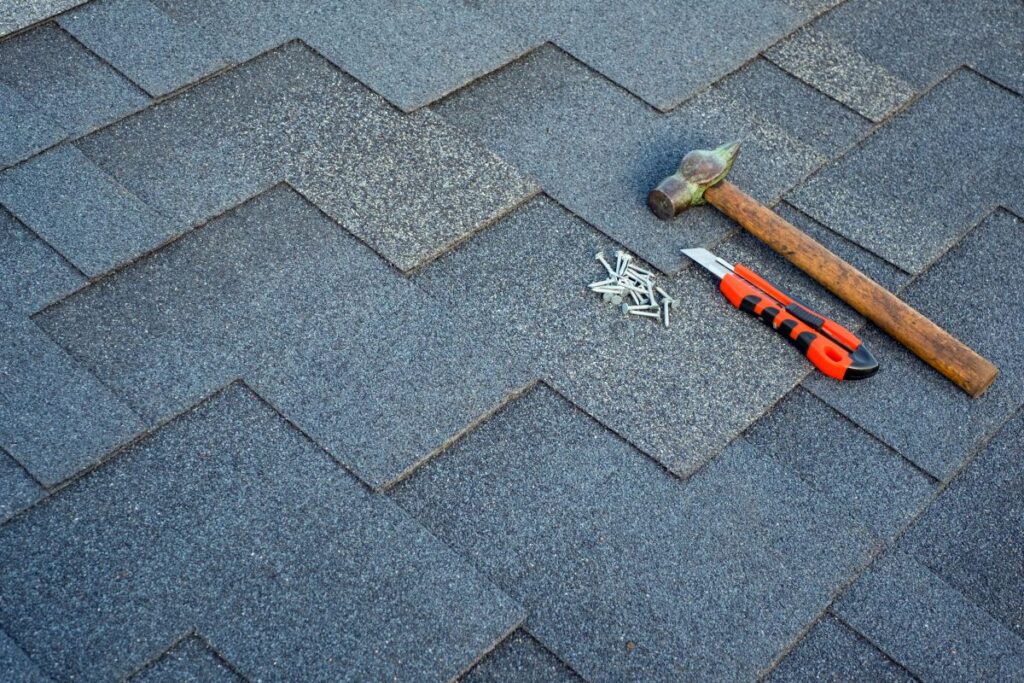 Gray asphalt roof shingles with a hammer, nails, and utility knife placed on top, suggesting roofing work or repair.