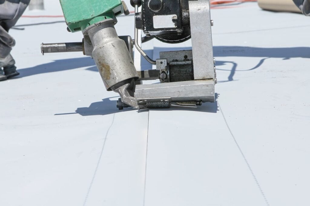 Close-up of a machine welding or sealing seams on a light-colored flat roof membrane, with a worker operating it.