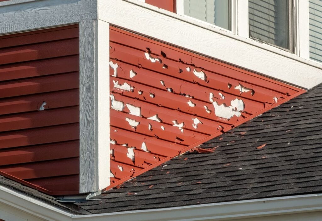 A section of red house siding with significant peeling and flaking white paint, next to white trim and black roof shingles, beneath a window.