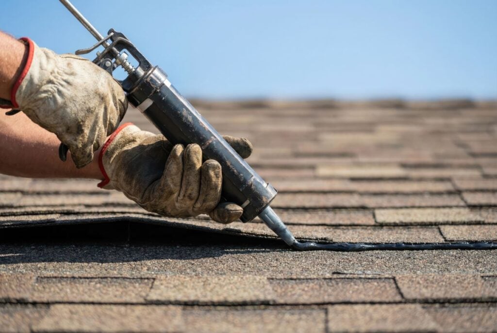 Gloved hands use a caulking gun to apply sealant along the seam of brown roof shingles under a clear blue sky.