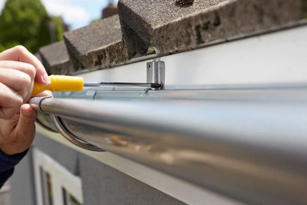 A person uses a screwdriver to attach a metal gutter to the roof edge of a house, securing a bracket under the shingles.