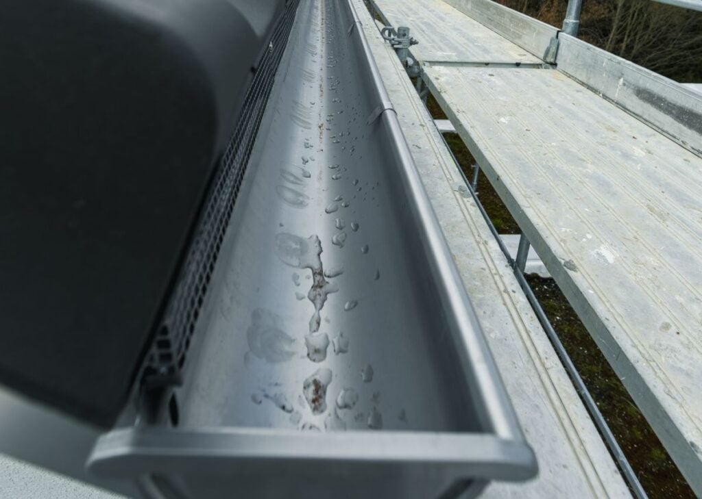 Close-up of a clean metal rain gutter with water droplets inside, attached to the edge of a roof next to a gray scaffolding platform.