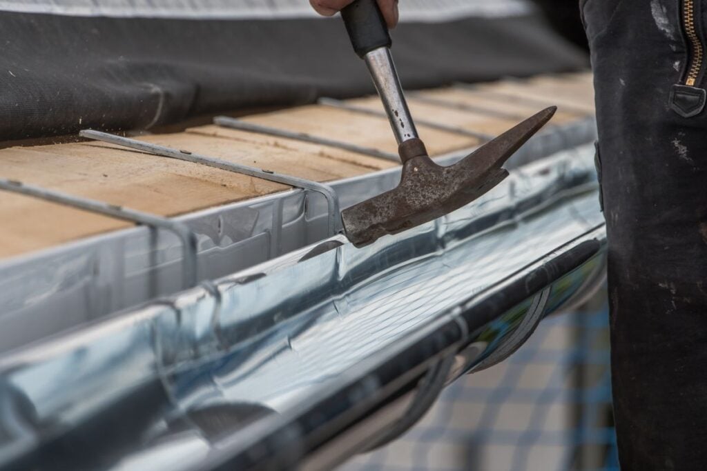 A person uses a hammer to secure metal flashing to a roof edge, with wooden planks and protective covering visible. The focus is on the hand holding the hammer near the metal gutter.