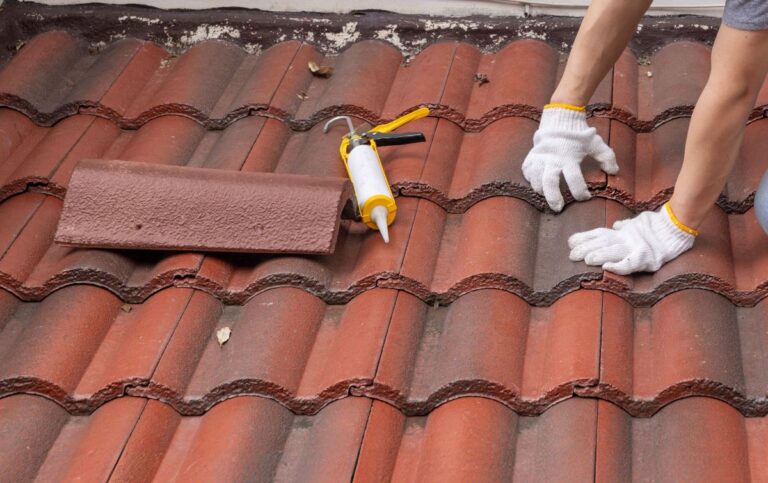 A person wearing white gloves repairs red roof tiles with a sealant, with a caulking gun and a loose tile nearby.