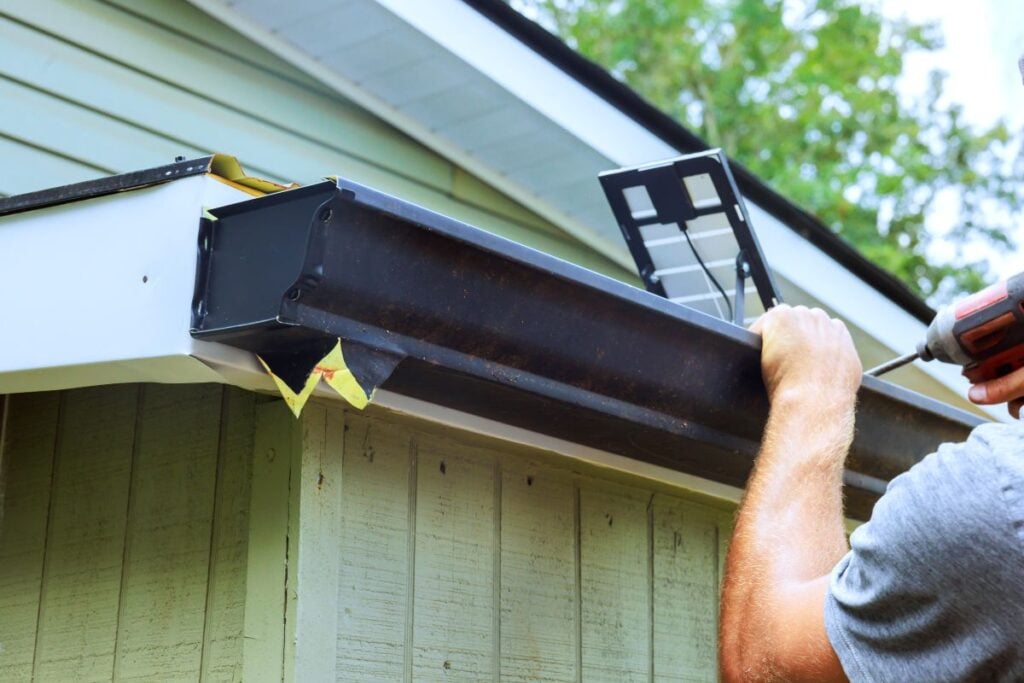 A person installs a rain gutter on a house, using a power drill. The house has light green siding, and trees are visible in the background.
