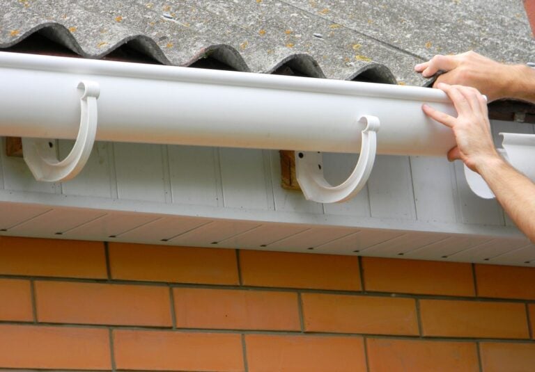 Two hands are installing a white plastic rain gutter onto the side of a house with orange brick walls and a tiled roof.