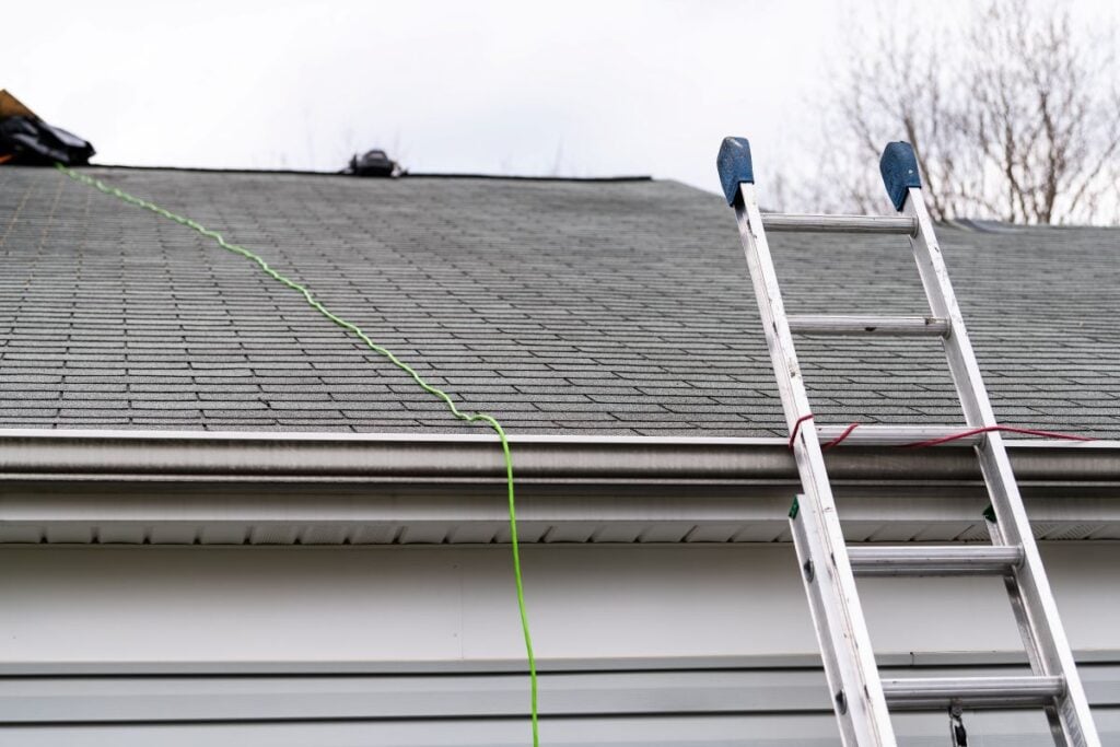 A silver ladder leans against a house roof. A green extension cord runs up the grey shingled roof, with some equipment visible near the roof peak. Trees without leaves are in the background.