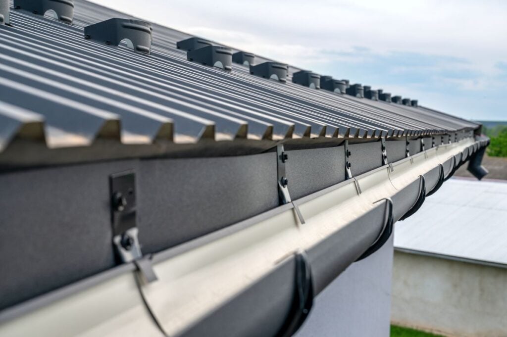Close-up of a metal roof with a gutter system attached, showing the edge of the roof, metal sheets, brackets, and fasteners. The background includes a clear sky and distant buildings.