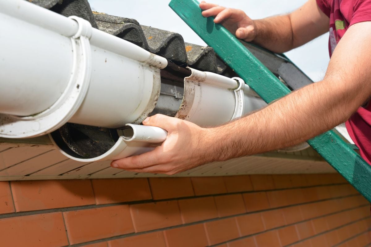 A person repairs a white rain gutter on a house, holding a section of the gutter while standing on a green ladder near a brick wall and roof tiles.