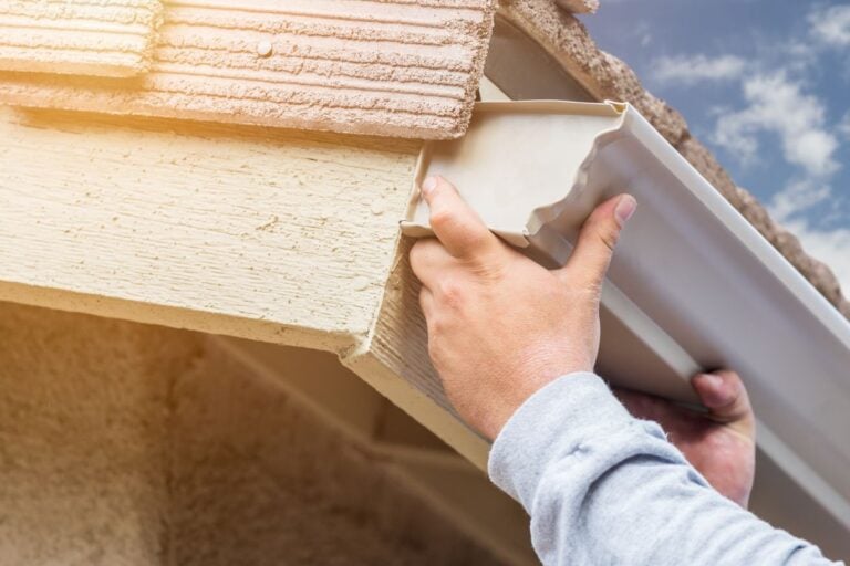 A person installs a white rain gutter on the edge of a house roof under a sunny sky.
