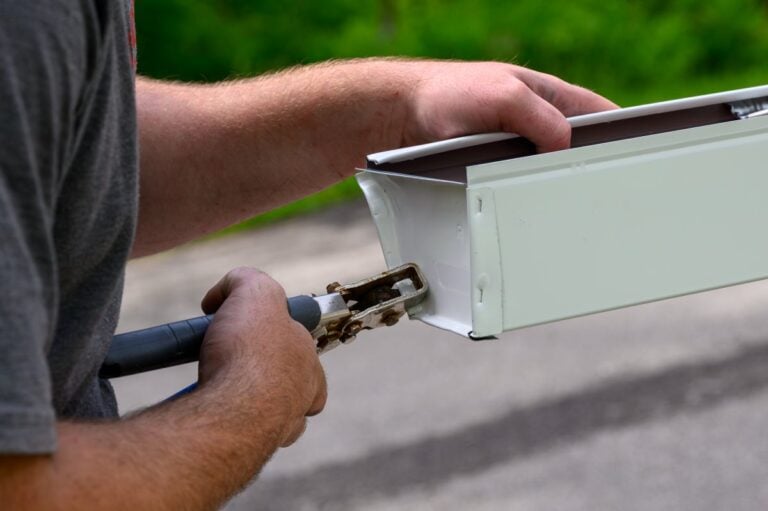 A person uses a handheld tool to crimp the end of a white metal gutter, preparing it for installation. The background is outdoors with green grass and pavement visible.