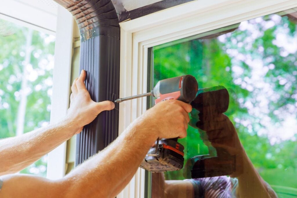 A person uses a power drill to attach a black downspout extension to the corner of a white window frame, with trees reflected in the window glass.