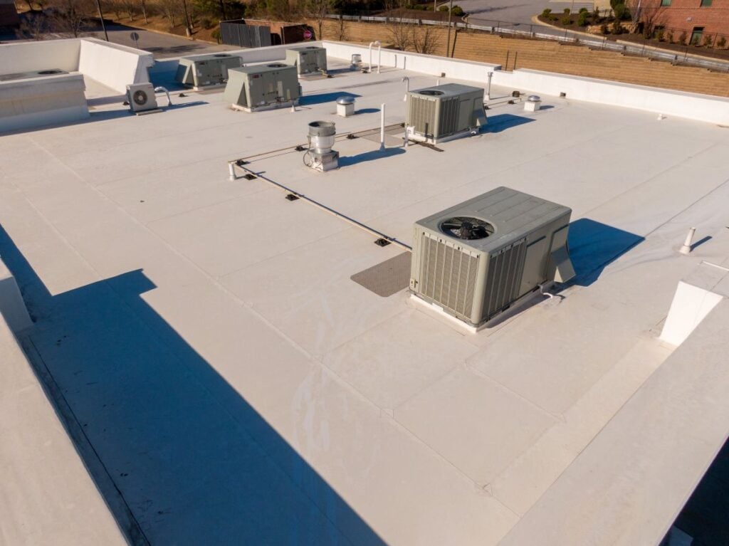 Aerial view of a flat white commercial building TPO roof with multiple HVAC units and vents, surrounded by a few roads and grassy areas.