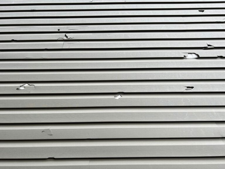 Close-up of light-colored vinyl siding with several cracks, tears, and holes across the surface, suggesting damage possibly caused by hail or impact. The siding panels are arranged horizontally.