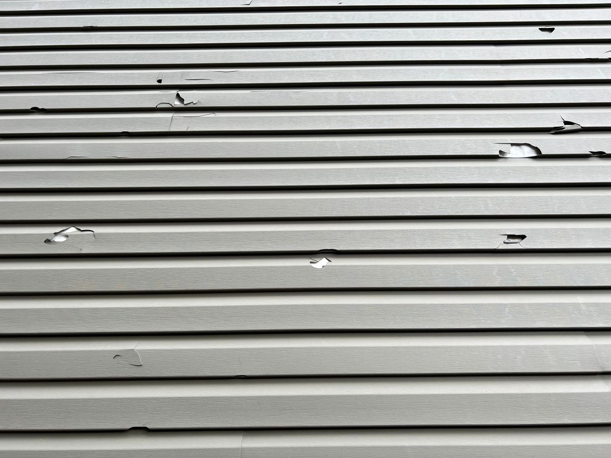 Close-up of light-colored vinyl siding with several cracks, tears, and holes across the surface, suggesting damage possibly caused by hail or impact. The siding panels are arranged horizontally.
