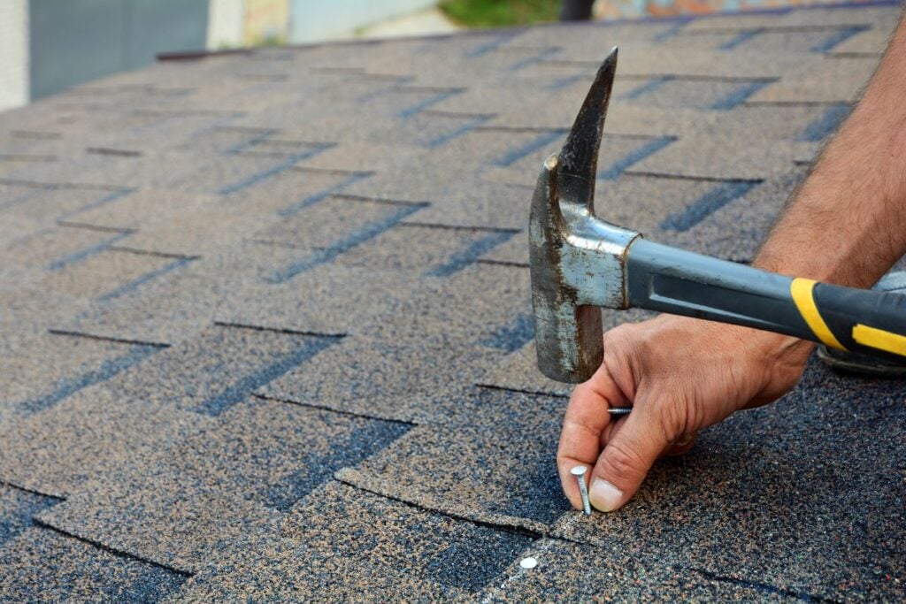 A person uses a hammer to nail down asphalt shingles on a roof, securing one shingle with a nail while holding it in place with their other hand.